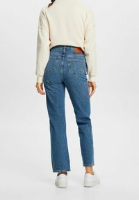 Vaqueros de denim azul de tiro alto con corte recto, con bolsillos traseros y un parche de cuero marrón. Combinados con un sudadera crema y zapatos blancos.