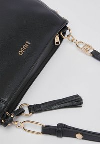 Sac à main en cuir noir avec une finition texturée, quincaillerie dorée, détail à pompon et bandoulière réglable. Comprend une fermeture éclair et un accent logo.