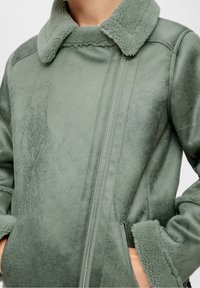 Groen-grijze shearling jas met diagonale rits, pluche kraag en manchetten, met handen deels in de ritszakken.