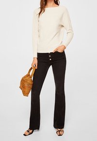 Off-White strukturierter Strickpullover mit weitem Ausschnitt, kombiniert mit schwarzen Schlagjeans mit Knopfverschluss vorne. Die Accessoires umfassen eine geflochtene Tasche.