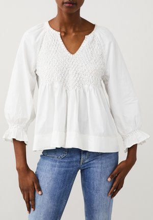 Femme portant un chemisier blanc texturé à manches longues avec encolure en V et poignets à volants, assorti à un jean en denim bleu.