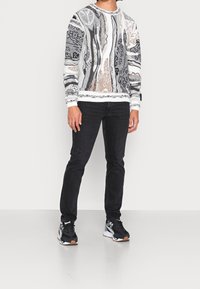 Pull avec motif abstrait en gris, blanc et marron ; col rond et poignets côtelés. Associé avec un jean noir et des baskets noires.