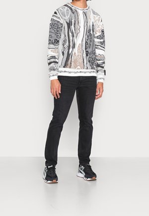 Pullover mit abstraktem Muster in Grau, Weiß und Braun; Rundhalsausschnitt und gerippte Bündchen. Kombiniert mit schwarzen Jeans und schwarzen Sneakers.