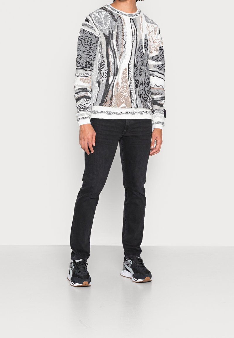 Pull avec motif abstrait en gris, blanc et marron ; col rond et poignets côtelés. Associé avec un jean noir et des baskets noires.