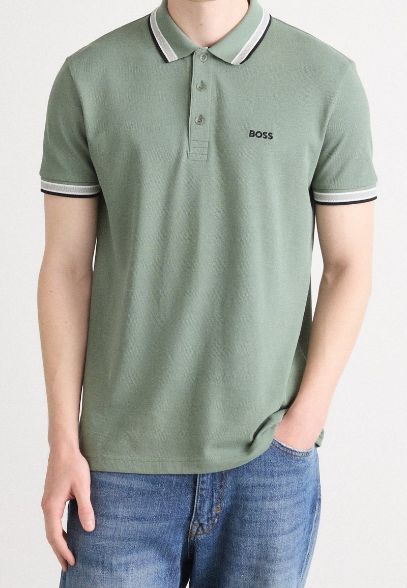Homme portant un polo vert clair avec un col et des manches à rayures blanches et noires, logo "BOSS" sur la poitrine, associé à un jean bleu.