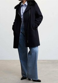 Femme portant un manteau noir avec un col en fourrure, une chemise bleu clair, un jean bleu à jambes larges et des chaussures noires à bout carré, debout sur un sol en béton.