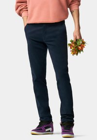 Pantalon bleu marine ajusté en tissu doux, associé à des baskets noires et violettes avec un accent blanc. Main tenant des fleurs orange.