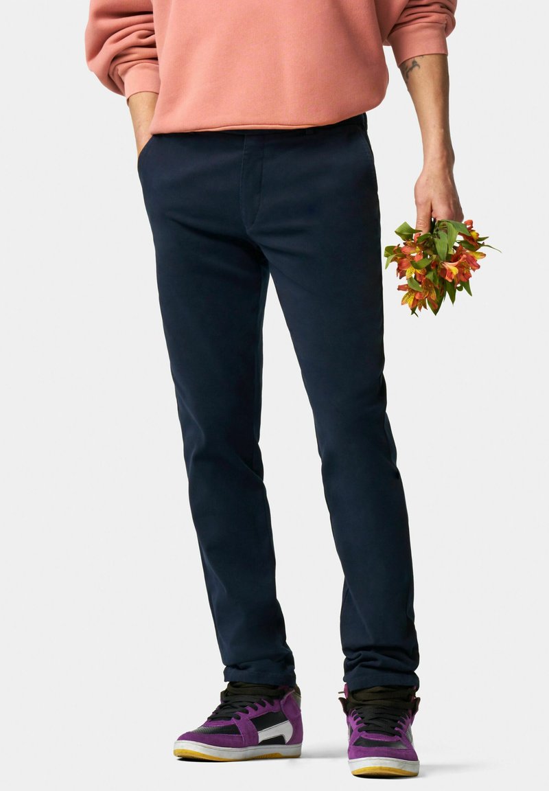 Pantalon bleu marine ajusté en tissu doux, associé à des baskets noires et violettes avec un accent blanc. Main tenant des fleurs orange.