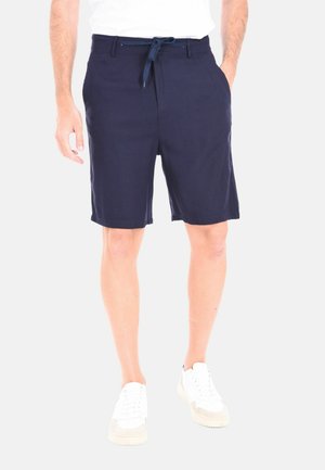 Uomo che indossa pantaloncini blu navy con coulisse e sneakers bianche, in piedi davanti a uno sfondo semplice.