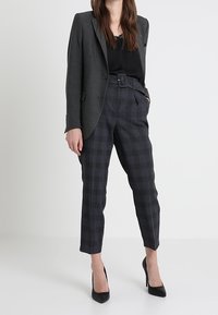 Femme portant un blazer gris foncé, un haut noir, un pantalon à carreaux taille haute avec ceinture, et des chaussures à talons noires, debout devant un fond blanc.