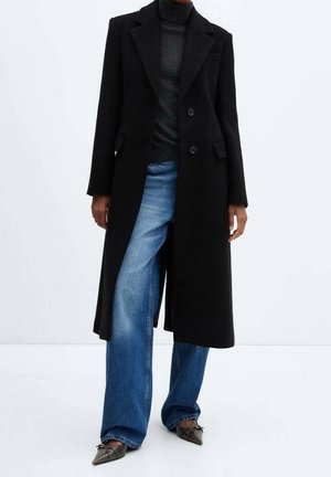 Person wearing a long black coat over a dark turtleneck and wide-leg blue jeans, paired with pointed-toe shoes with bows.
