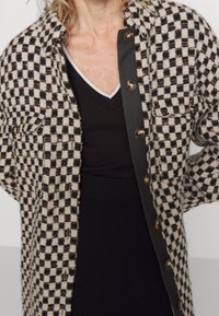 Geruit jasje in beige en zwarte stof, met grote knopen, twee zakken aan de voorkant en een losse pasvorm met een contrasterende kraag.