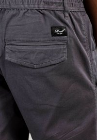 Pantalones de algodón grises con cintura elástica, presentan un bolsillo trasero y un diseño cosido. Una etiqueta oscura con texto blanco es visible.