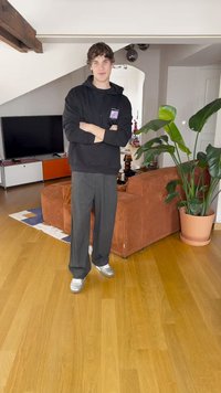 Melnā kapuce, pelēkie platās bikses un sudraba apavi. Interjers ar koka grīdu, brūnu ribotu dīvānu un podu ar lapu augu.