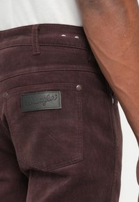 Pantalone od kordura u dubokoj burgundijskoj nijansi s vidljivim teksturiranim naborima, sa crnom kožnom oznakom i metalnim gumbima na džepovima.