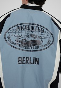 Personne portant une veste bleue et noire avec l'inscription "Prohibited Racing Club" et un motif de voiture imprimé au dos, avec "Berlin" en dessous.