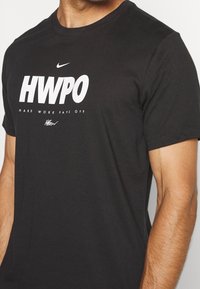 Camiseta de algodón negra con cuello redondo. Presenta texto blanco: "HWPO HARD WORK PAYS OFF" y un logo de Nike arriba. Manga corta, ajuste regular.