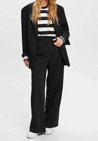 Černý oversize blazer přes černo-bílou pruhovanou košili, spárovaný s širokými černými kalhotami a bílými teniskami. Hladká tkanina, precizní střih.