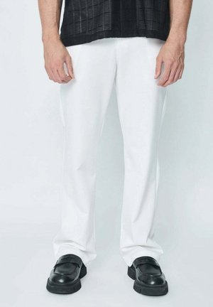 Homme portant un pantalon blanc droit et des chaussures noires sans lacets, debout devant un fond clair uni.