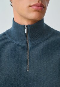 Maglione lavorato a maglia con collo alto di colore blu scuro. Presenta una zip argentata e una trama a costine verticali per un tocco di dettaglio.