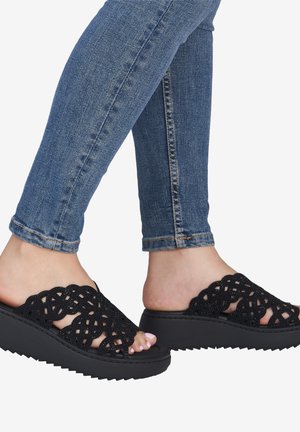 Pieds portant un jean bleu et des sandales compensées noires à bouts ouverts avec des brides tissées et une légère semelle plateforme.
