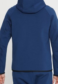 Sweat à capuche bleu marine à texture lisse, doté d'une coupe ajustée, d'une capuche et d'un accent noir à l'ourlet. Vue arrière montrant des manches raglan.