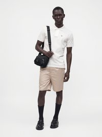 Homme portant un polo blanc, un short beige, des longues chaussettes noires, des chaussures noires et portant un sac bandoulière noir sur un fond uni.