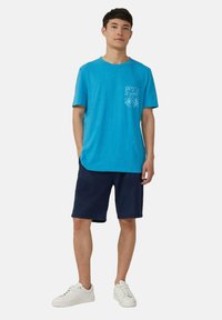 Blauwe katoenen T-shirt met een ronde hals en een borstzak met een witte bloemenprint, gecombineerd met marineblauwe shorts en witte sneakers.