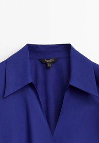 Lila blus med spetsig krage och slät yta. Har en logotypetikett synlig inuti kragen.