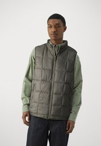 Grå quilted vest med høy krage og glidelås, sammen med en grønn langermet skjorte. Merkeverdige teksturerte mønstre på vesten.