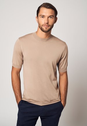 Mann mit kurzen braunen Haaren und Bart, trägt ein beiges Kurzarmhemd und dunkle Hosen, steht mit den Händen in den Taschen vor einem schlichten Hintergrund.