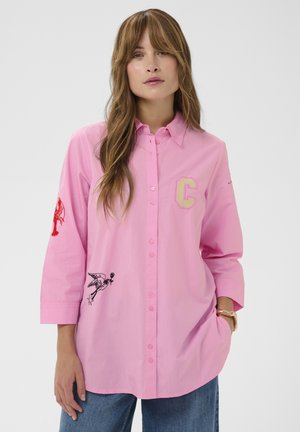 Hosszú hajú nő, rózsaszín, gombos ingben, amelyen sárga "C" betű, az ujjon piros homár, valamint a szegély közelében fekete madár ábrázolás található.