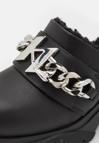 Chaussure noire avec une tige en cuir synthétique, dotée d'un accent en chaîne argentée proéminent et d'un détail logo. Surface texturée et semelle robuste.