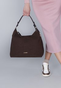 Sac à main en daim marron de forme distinctive, avec un logo doré, complété par des anses en cuir noir et une fermeture éclair.