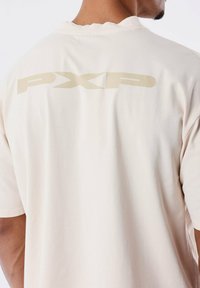 Camiseta beige con cuello redondo y mangas cortas. Presenta un gran logo "PXP" de color claro impreso en la parte superior de la espalda.
