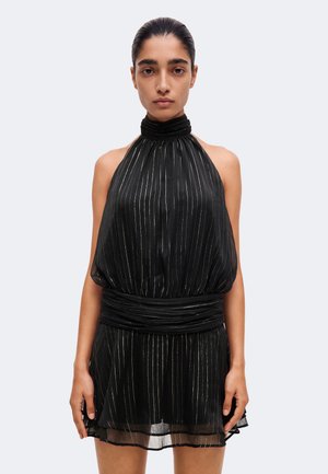 Une femme se tient face à l’avant, vêtue d’une robe noire sans manches à nouer autour du cou, avec de fines rayures verticales et une taille froncée.
