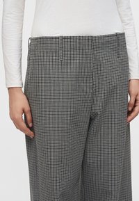 Personne portant un pantalon à carreaux gris et noirs et une chemise blanche à manches longues, mains posées près des hanches, sur un fond uni.