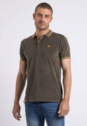 Homme portant un polo marron avec un bord orange et un jean bleu, debout devant un fond clair uni.