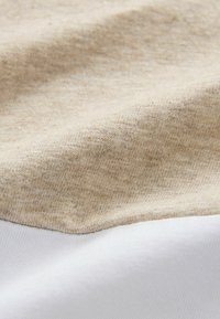 Textielstof met een beige, gestructureerd oppervlak en een gladde witte onderkant, die een zachte, geweven kwaliteit en contrasterende kleuren toont.