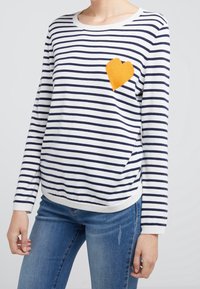 Pull tricot rayé blanc et marine avec un patch cœur jaune sur le côté gauche. Doté d'un col rond et de manches longues.