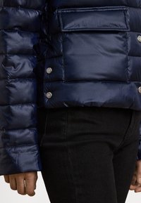 Μπουφάν navy μπλε με γυαλιστερό φινίρισμα, με οριζόντιες ραμμένες γραμμές, κουμπιά snap και τσέπη στο στήθος. Μαύρο τζιν εμφανές από κάτω.