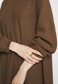 Brun ribbad stickad tröja med uppvikt ärm; en hand prydd med en guldig texturerad ring vilar på tyget. Mysig, avslappnad passform.