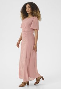 Pink maxi dress in lightweight fabric with short, puffed sleeves and gathered detailing at the waist. Paired with brown block-heeled sandals.