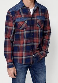 Homme portant une chemise à carreaux rouge et bleu boutonnée avec des détails en denim sur les poches et les épaules, associée à un jean bleu.