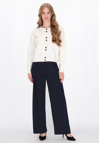 Crème cardigan met ronde hals, zwarte knopen en een klein zakje. Gecombineerd met marineblauwe wijde broek en zwarte hakken. Glad tekstuur.