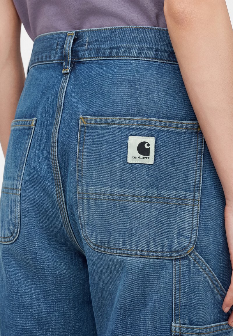 Close-up of a person wearing blue denim jeans with large back pockets, one featuring a Carhartt logo patch.