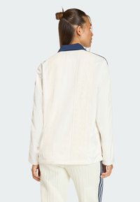 Biela prešívaná bunda s textúrovaným vertikálnym vzorom, tmavomodrým golierom a dlhými rukávmi. Zvýraznená voľným strihom, kombinovaná s krémovými nohavicami.