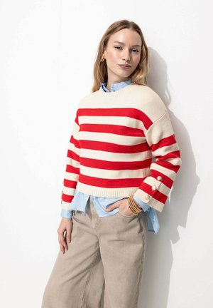 Pull rayé rouge et crème avec des manches longues et des accents de boutons ; porté sur une chemise bleu clair. Le modèle porte un pantalon large beige.