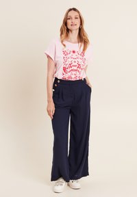 Roze grafisch T-shirt met rood bloemenmotief en tekst, gecombineerd met marineblauwe wijde broek met gouden knopen. Witte sneakers gedragen.