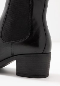 Czarne skórzane botki za kostkę z teksturowaną elastyczną boczną wstawką, zaokrąglonym noskiem oraz niskim blokowym obcasem z ryflowanym detalem.